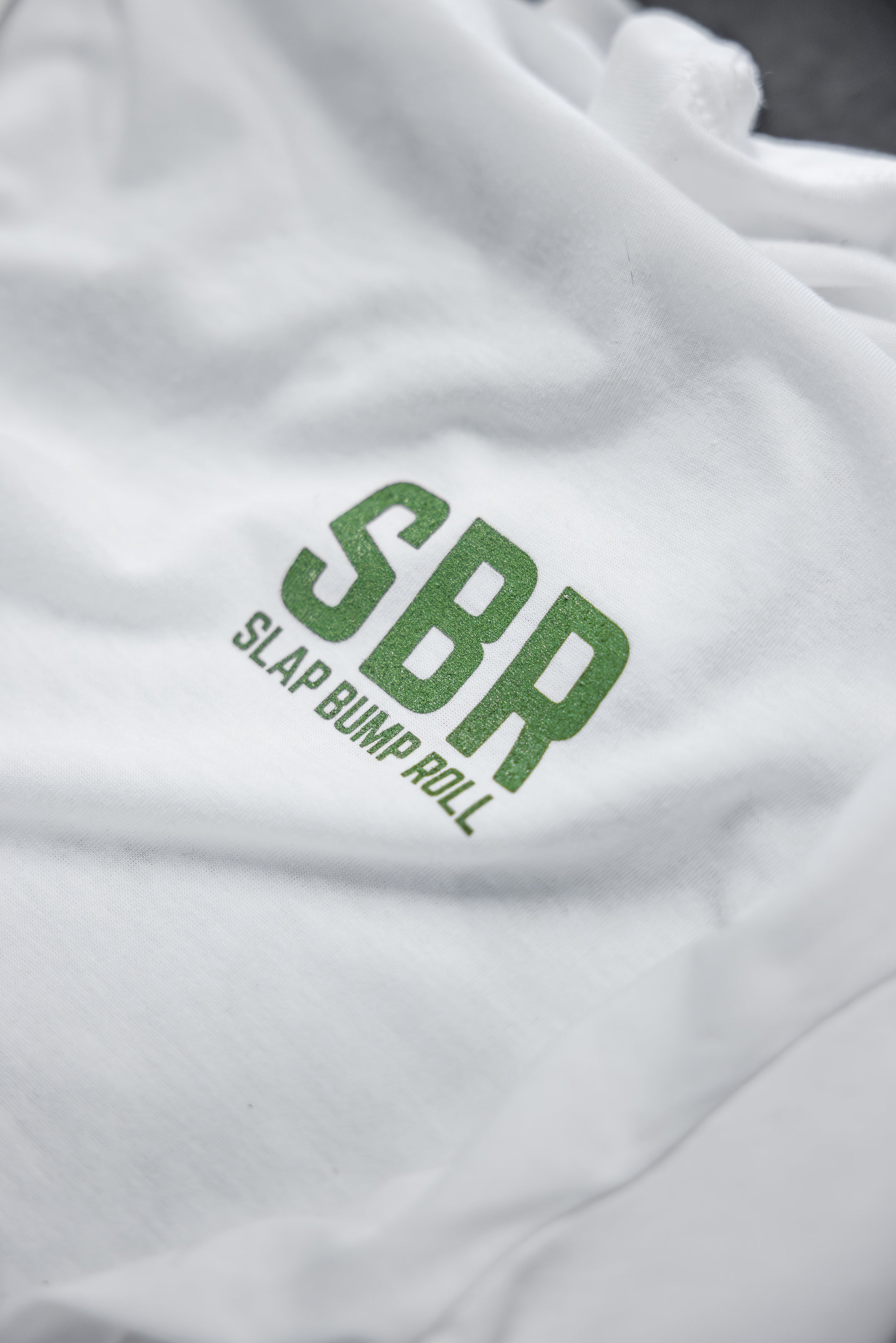 Close-up of the green SBR Slap Bump Roll wordmark on the chest of the soft white tri-blend jiu jitsu tee