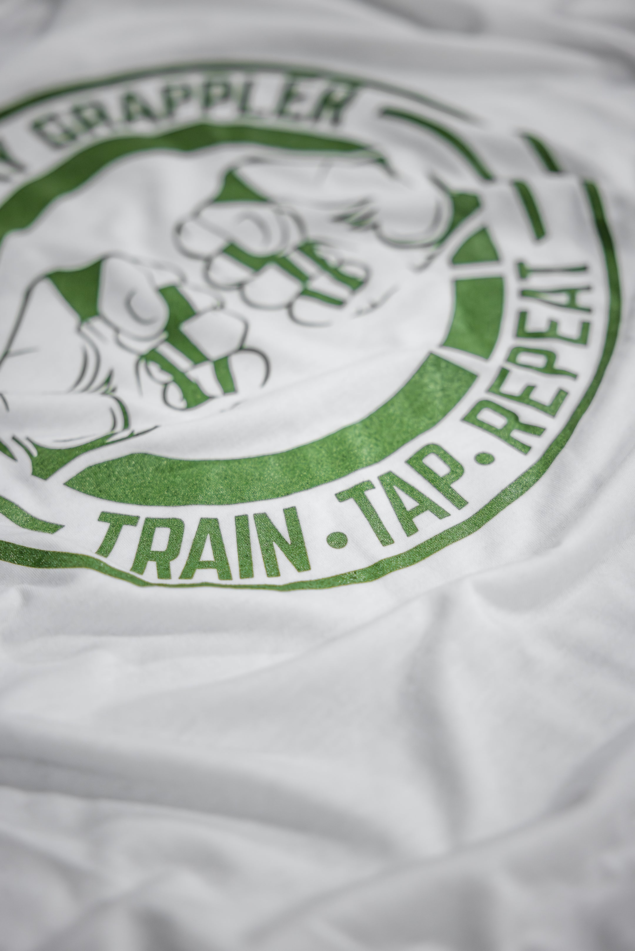 Detail of the green TRAIN TAP REPEAT slogan circling the fist-bump logo on the Everyday Grappler BJJ tee
