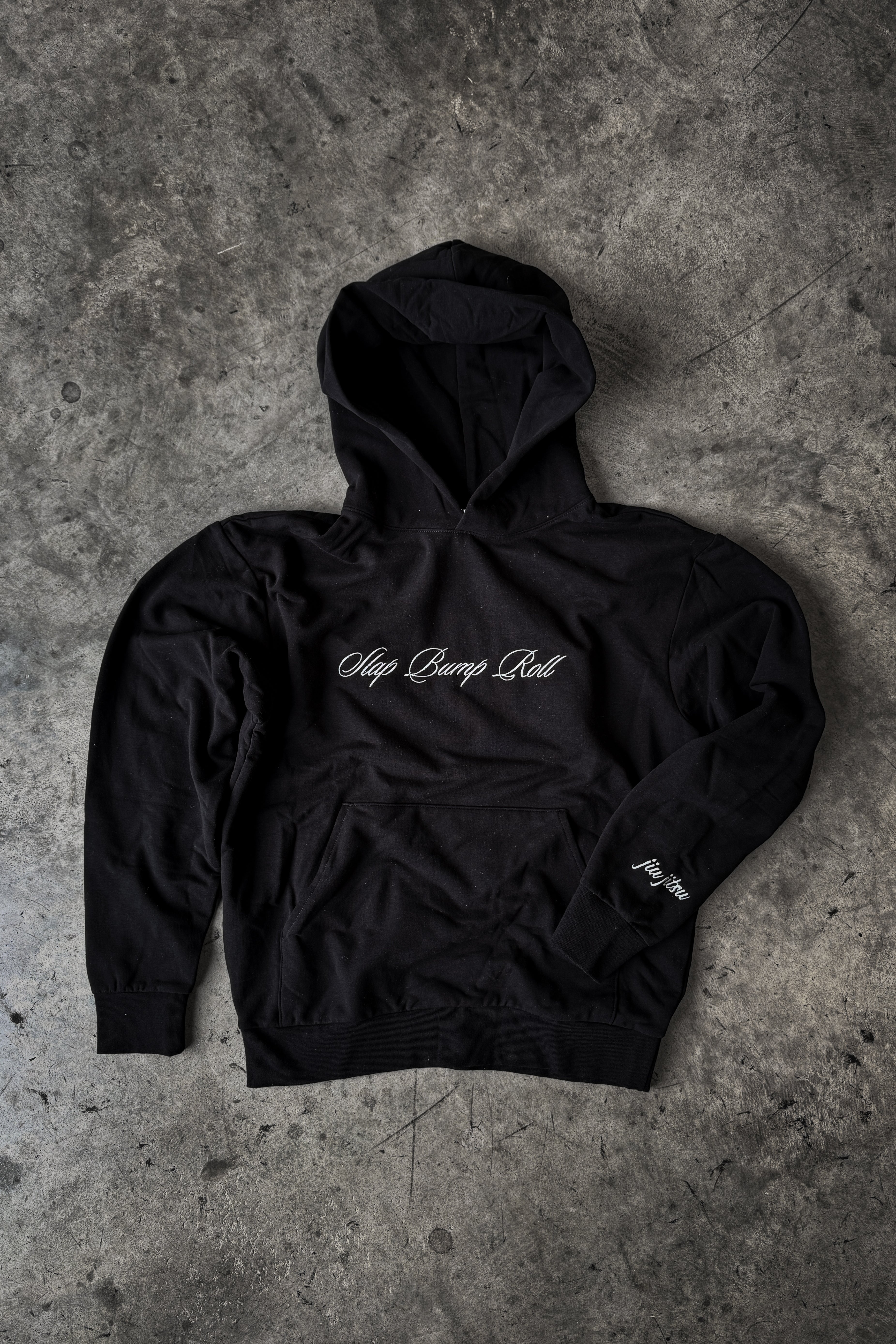 SBR Black Butter Hoodie flat lay on concrete, premium heavyweight BJJ lifestyle hoodie by Slap Bump Roll