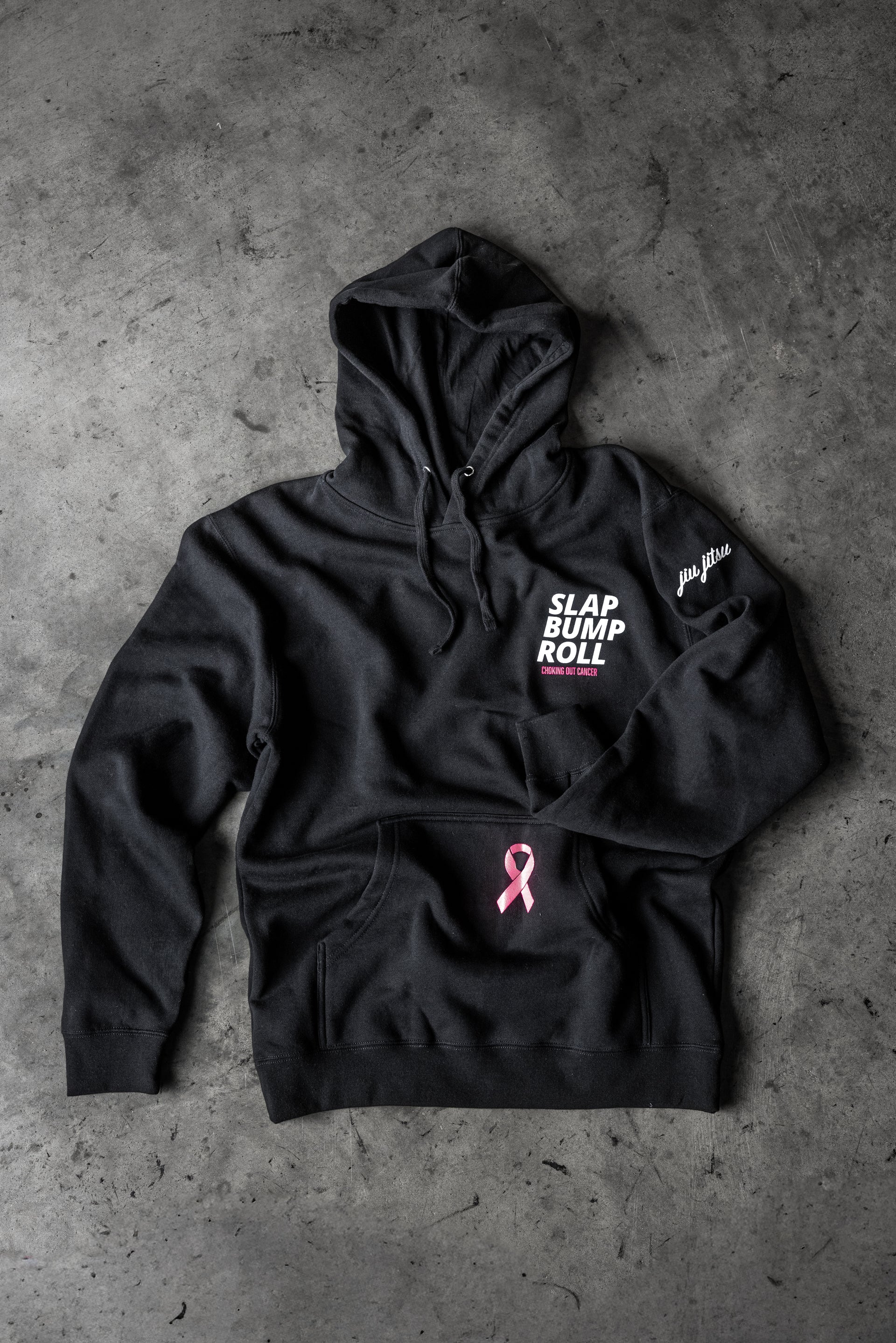 SBR Choking Out Cancer Hoodie in black, flat lay with SLAP BUMP ROLL chest print and pink awareness ribbon on pocket
