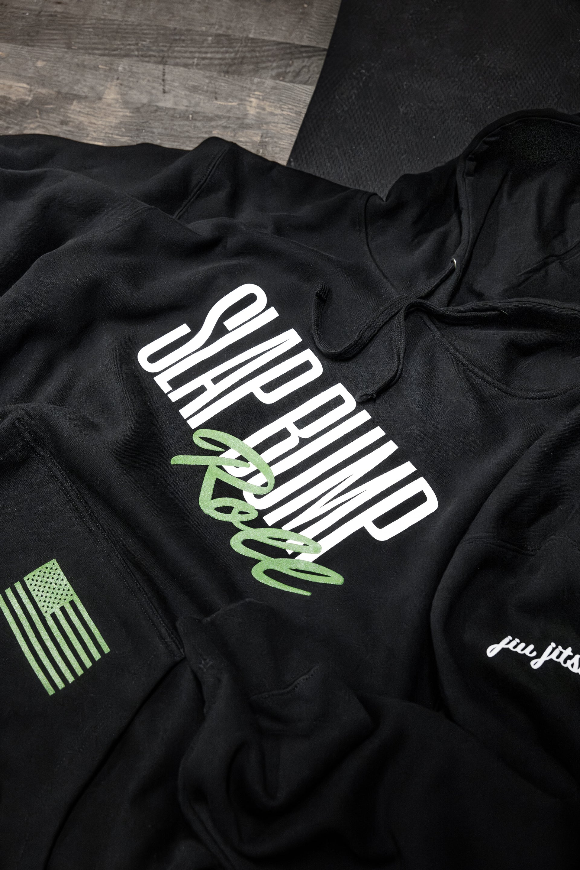 Black jiu jitsu hoodie detail showing the SLAP BUMP ROLL print, green American flag on the cuff, and jiu jitsu script