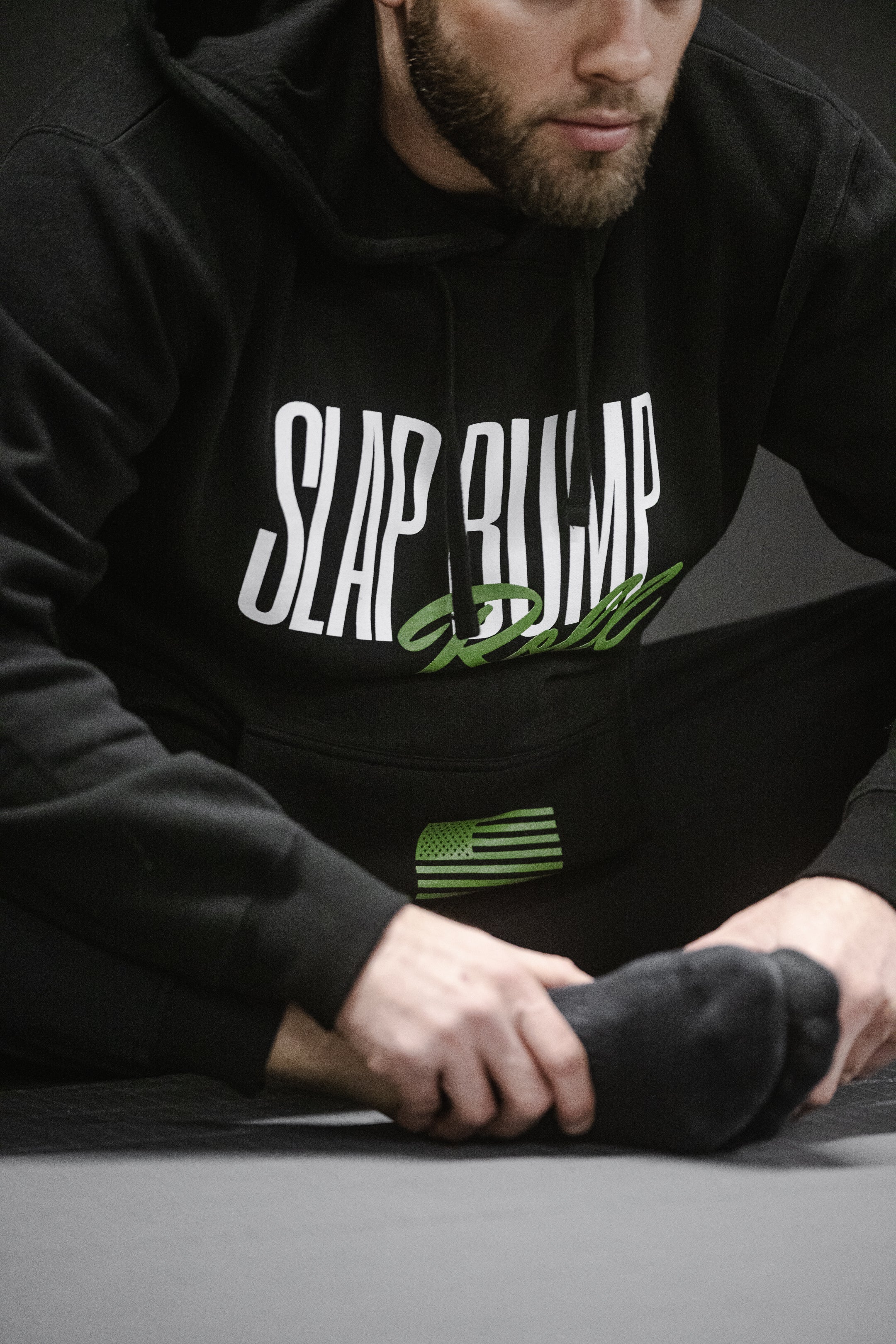 Grappler kneeling on the mat in the black Slap Bump Roll BJJ hoodie with white SLAP BUMP ROLL wordmark and hand wraps